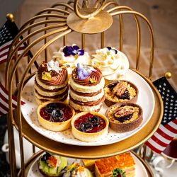 A three-tiered afternoon tea stand featuring an assortment of elegant pastries and savory bites, including layered cakes topped with edible flowers, fruit tarts, pecan tartlets, tea sandwiches, and whipped cream desserts. Small American flags frame the display, creating a festive patriotic presentation.