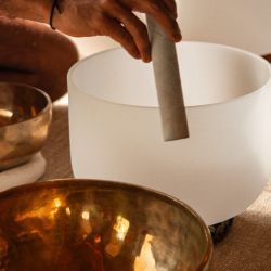 Hand holding a mallet above a white crystal singing bowl, surrounded by glowing brass bowls in a warm, serene wellness setting.