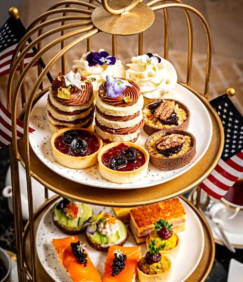 A patriotic afternoon tea display presented on a gold two-tier stand adorned with small American flags. The top tier features an assortment of elegant pastries, including layered cakes topped with edible flowers, berry tarts, and pecan tartlets. The lower tier showcases savory bites such as smoked salmon canapés with caviar, cucumber sandwiches, and miniature tartlets garnished with herbs. The refined presentation evokes a celebratory America 250-themed tea experience.